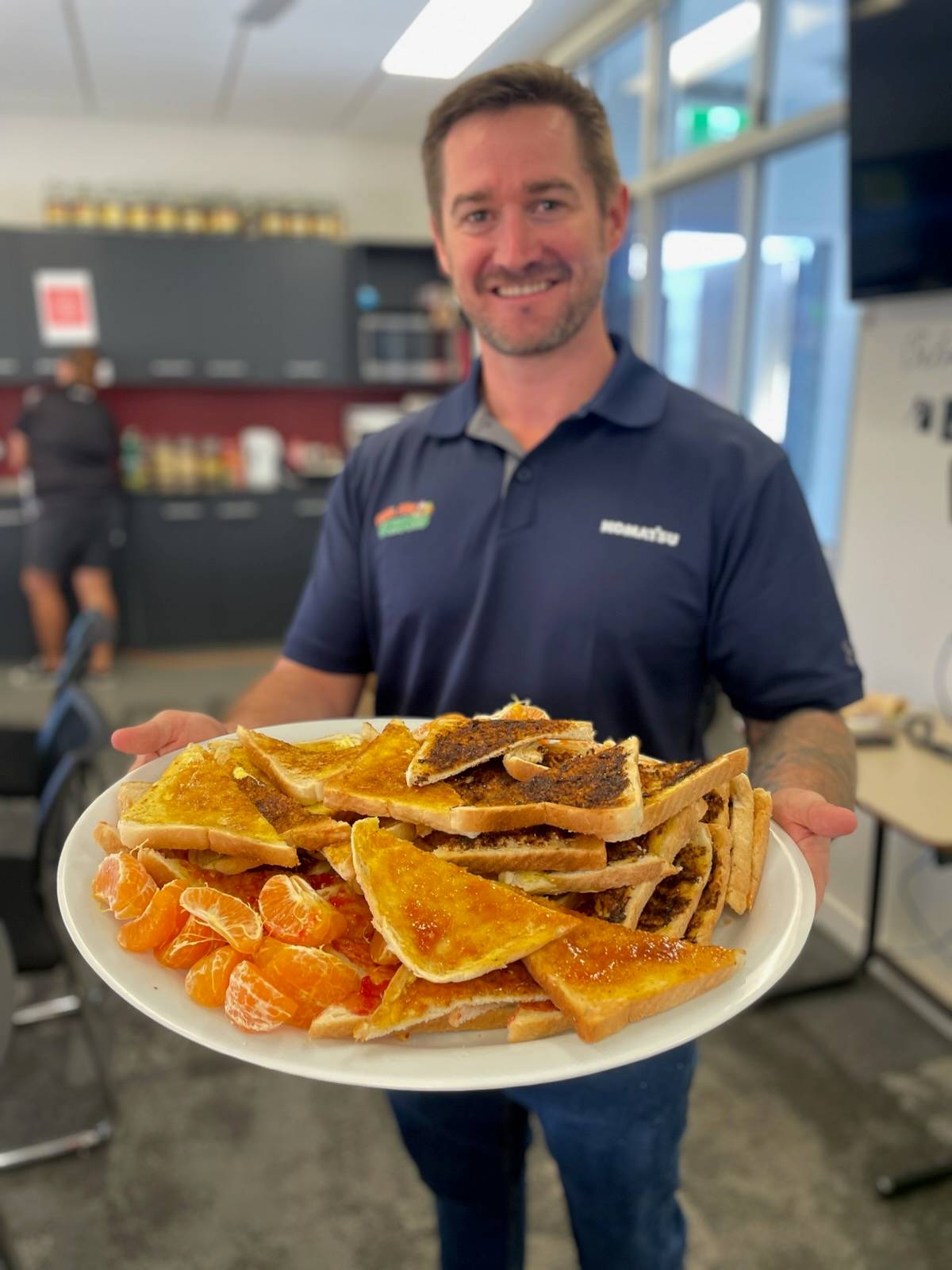 An individual wearing a blue Komatsu and Live Your Dream polo, smiling and holding a large plate of assorted foods.