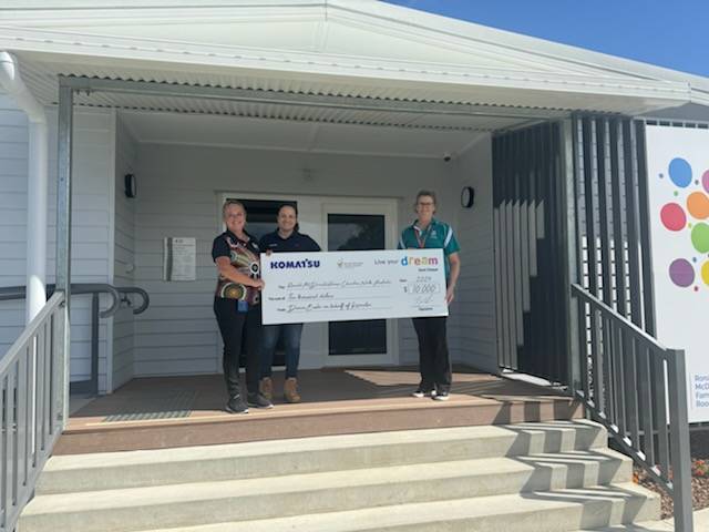 Three individuals standing in front of a building entrance, holding an oversized ceremonial check from Komatsu.