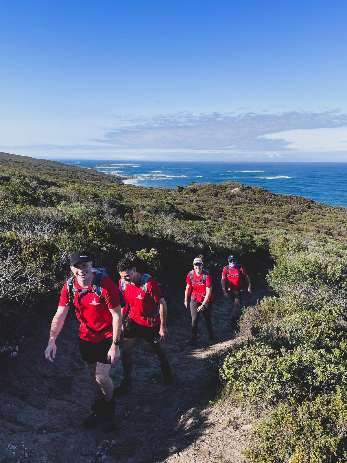Four individuals wearing red shirts hiking on a coastal trail with lush greenery and a view of the ocean in the background.
