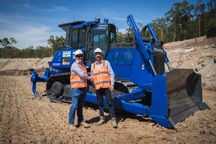 Two workers in safety vests and helmets stand in front of a Komatsu bulldozer at a construction site.