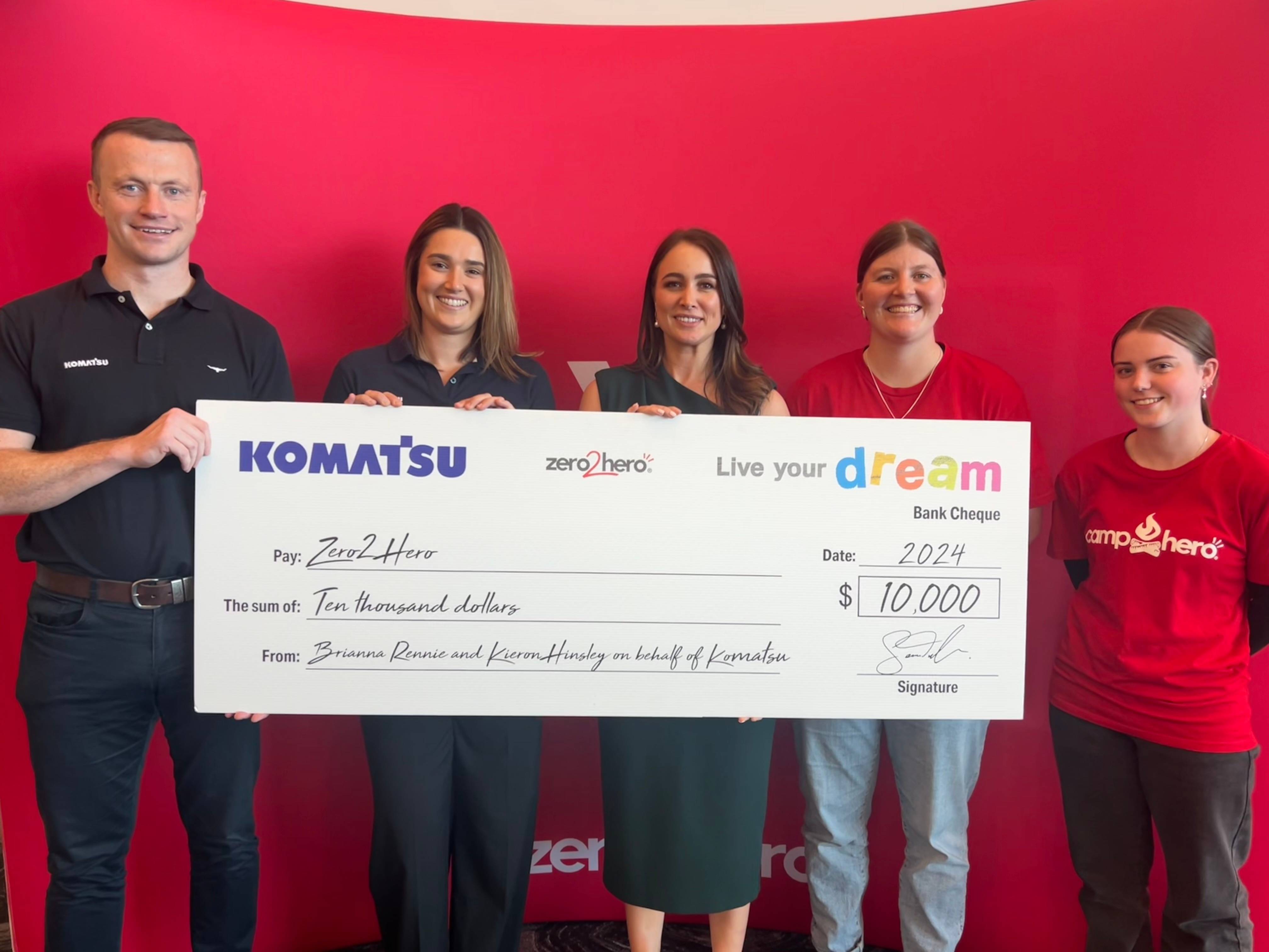 Five individuals standing in front of a red wall with the Komatsu logo, holding an oversized promotional check from Komatsu to Zero2Hero for ten thousand dollars.