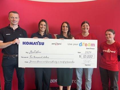 Five individuals standing in front of a red wall with the Komatsu logo, holding an oversized promotional check from Komatsu to Zero2Hero for ten thousand dollars.