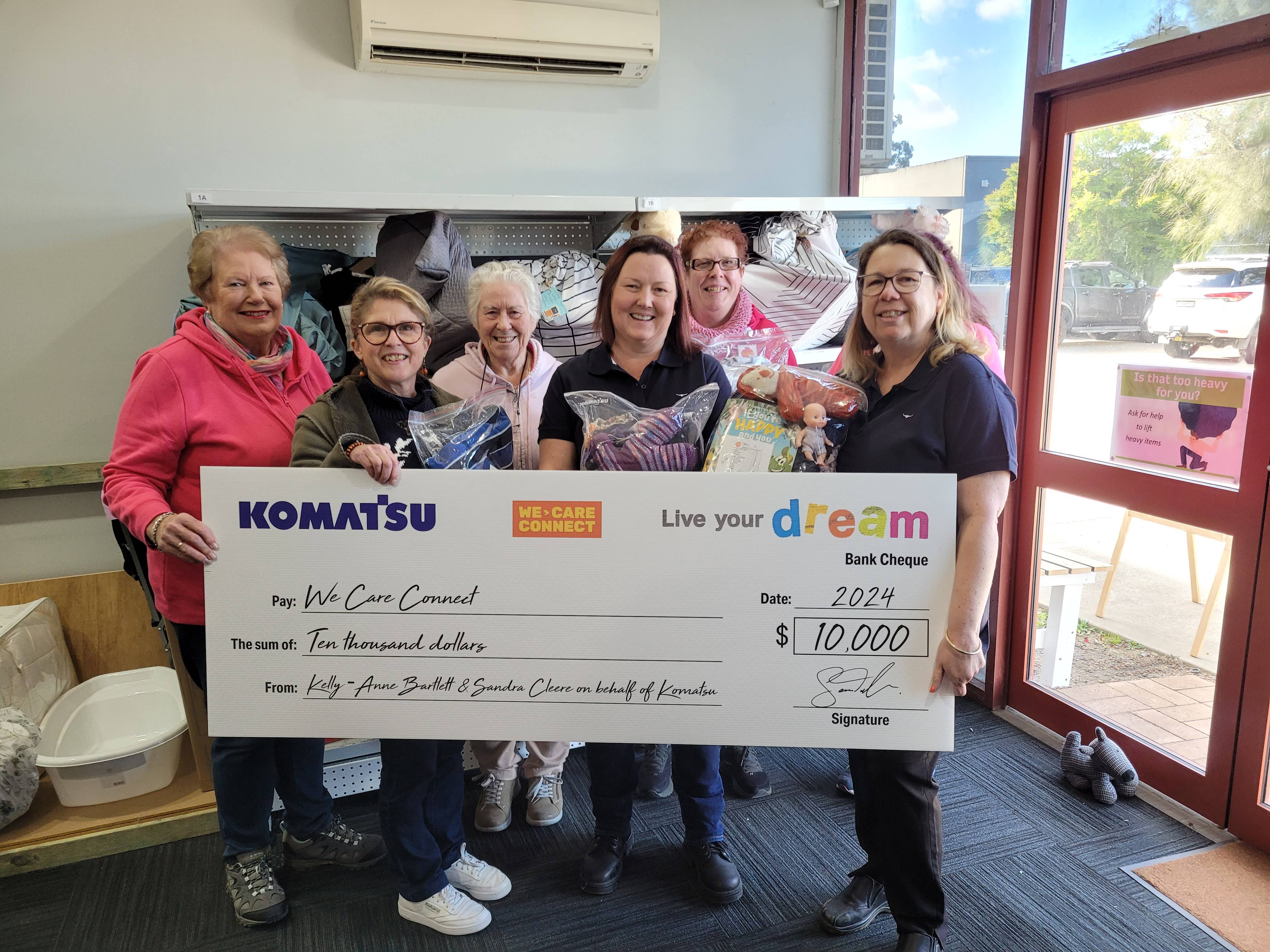 Five people stand together smiling as they present a large promotional check from Komatsu to We Care Connect.