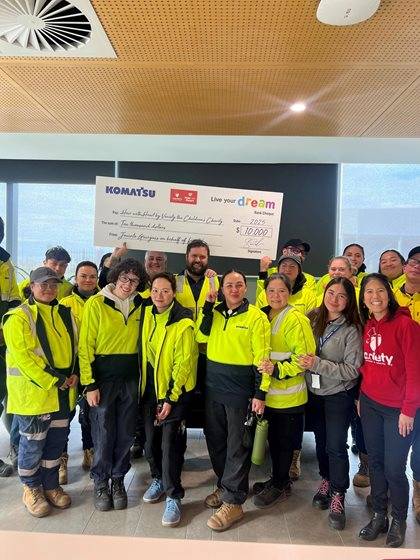 Group of workers in yellow jackets standing with a person holding a large check from Komatsu for $10,000, under a banner reading 'Live Your Dream.'