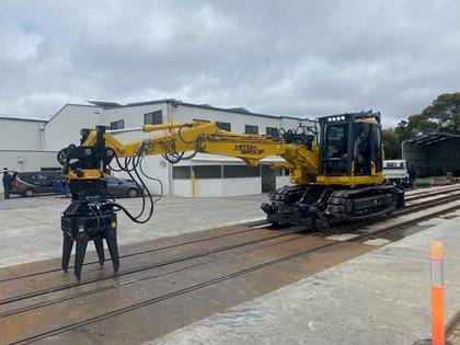 A Komatsu construction rail excavator equipped with a grapple attachment, parked on railway tracks in an industrial yard.