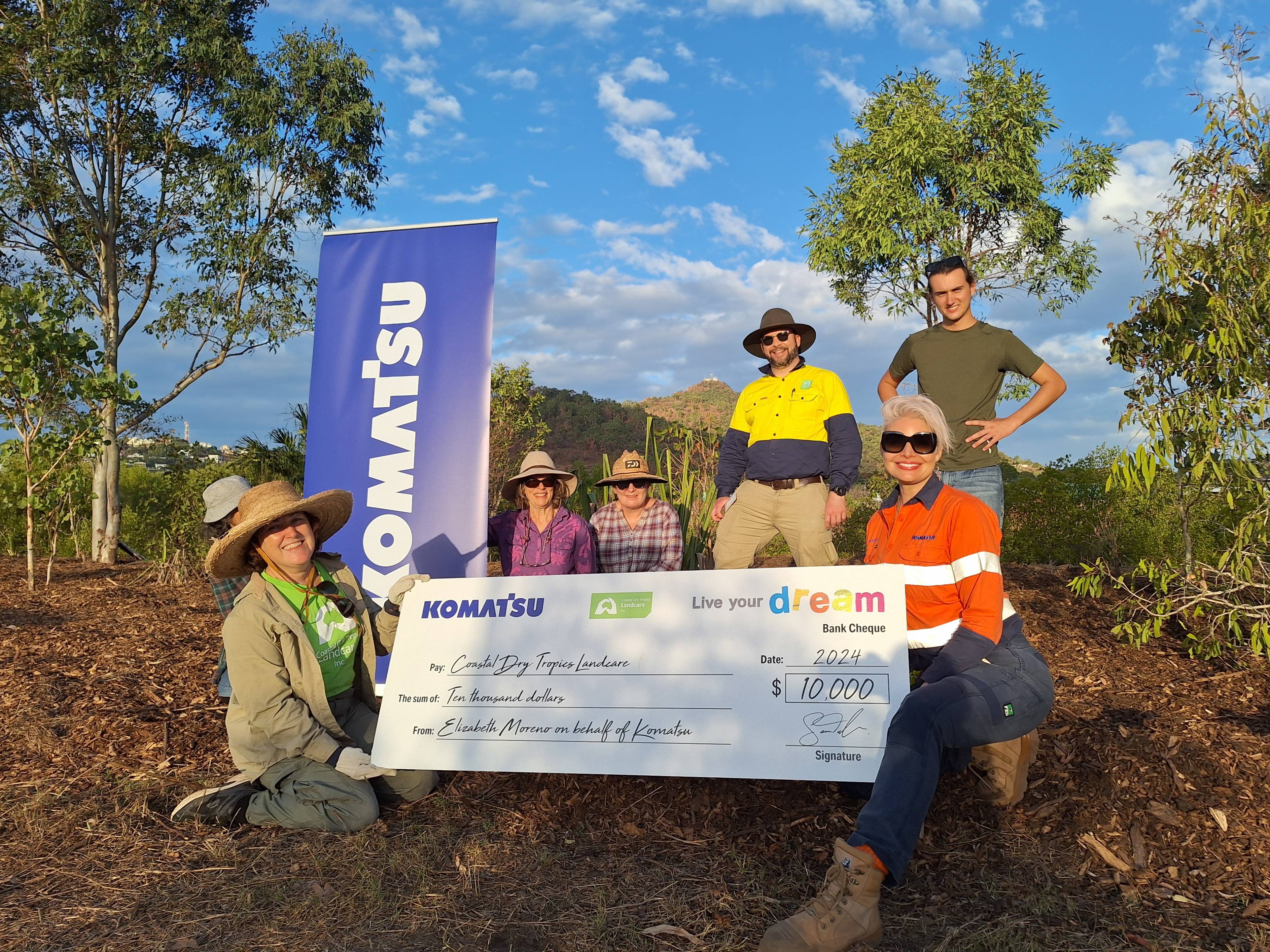 Group of five people posing outdoors with a large check from Komatsu. They are surrounded by natural scenery under a clear sky.