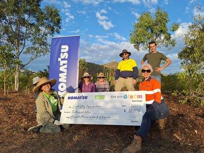 Group of five people posing outdoors with a large check from Komatsu. They are surrounded by natural scenery under a clear sky.