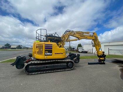 A Komatsu PC138US excavator parked on an asphalt surface, with commercial buildings under a cloudy sky in the background.