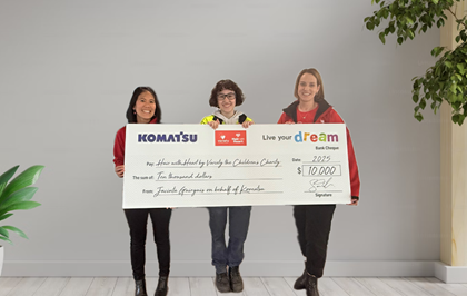 Three individuals hold a large ceremonial check from Komatsu to Live Your Dream for $10,000 addressed to Hair with Heart by Variety the Children's Charity.