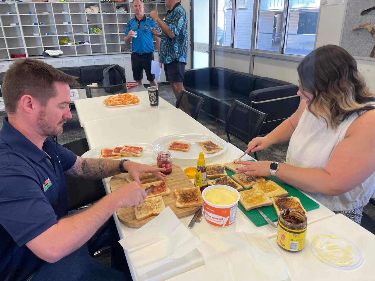 Two people are sitting down at a table and applying spread onto toasts.