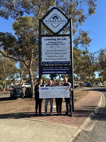 Four individuals holding a large ceremonial check in front of the Wirreanda Secondary School sign under a clear blue sky.