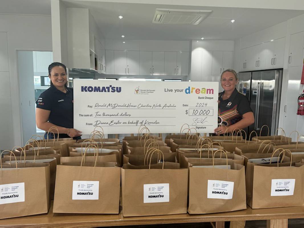 Two individuals stand behind a counter, displaying a large Komatsu check for $10,000, surrounded by brown paper bags arranged on the countertop, inside a bright kitchen area.