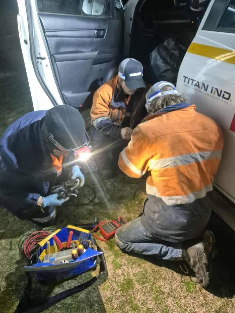 Three workers in high-visibility jackets and helmets are using welding equipment on a metal bar next to a vehicle at night. One worker is welding while the others observe, surrounded by various tools and equipment.