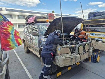 Two individuals examine the open engine bay of a heavily decorated rally car in a parking lot. The car is covered in stickers and equipped with additional exterior gear.