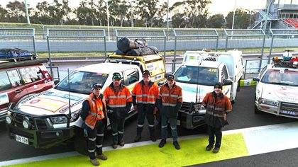 Five individuals in high-visibility vests standing in front of two rally support vehicles on a racing circuit.