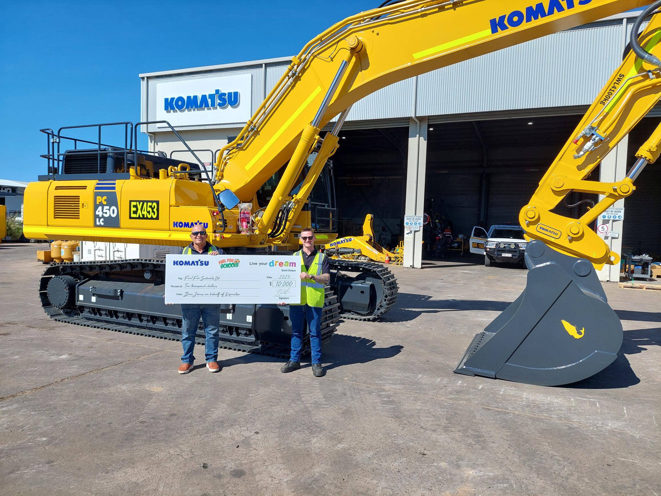 Two individuals standing standing in front of a yellow Komatsu excavator, holding a large check for ten thousand dollars, made out to Fuel for Schools.