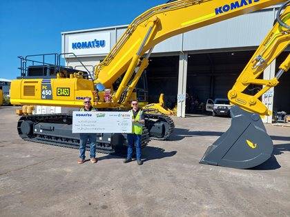 Two individuals standing standing in front of a yellow Komatsu excavator, holding a large check for ten thousand dollars, made out to Fuel for Schools.