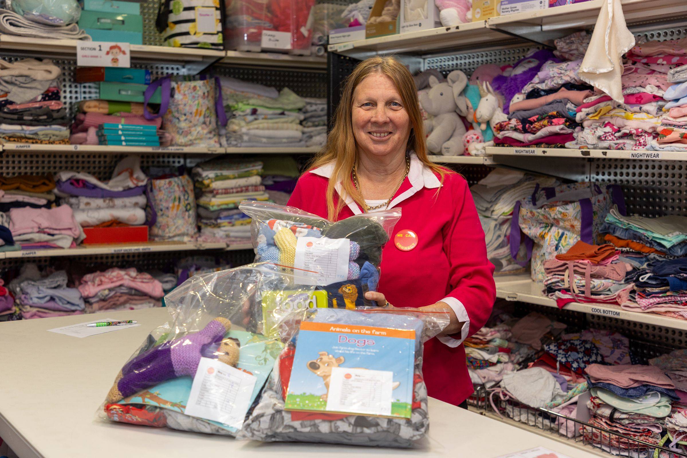 A woman smiling, standing behind a counter with various baby products including clothing and toys.
