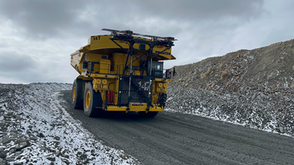 A large yellow Komatsu dump truck driving on a gravel road with snow-dusted rocky terrain in the background.