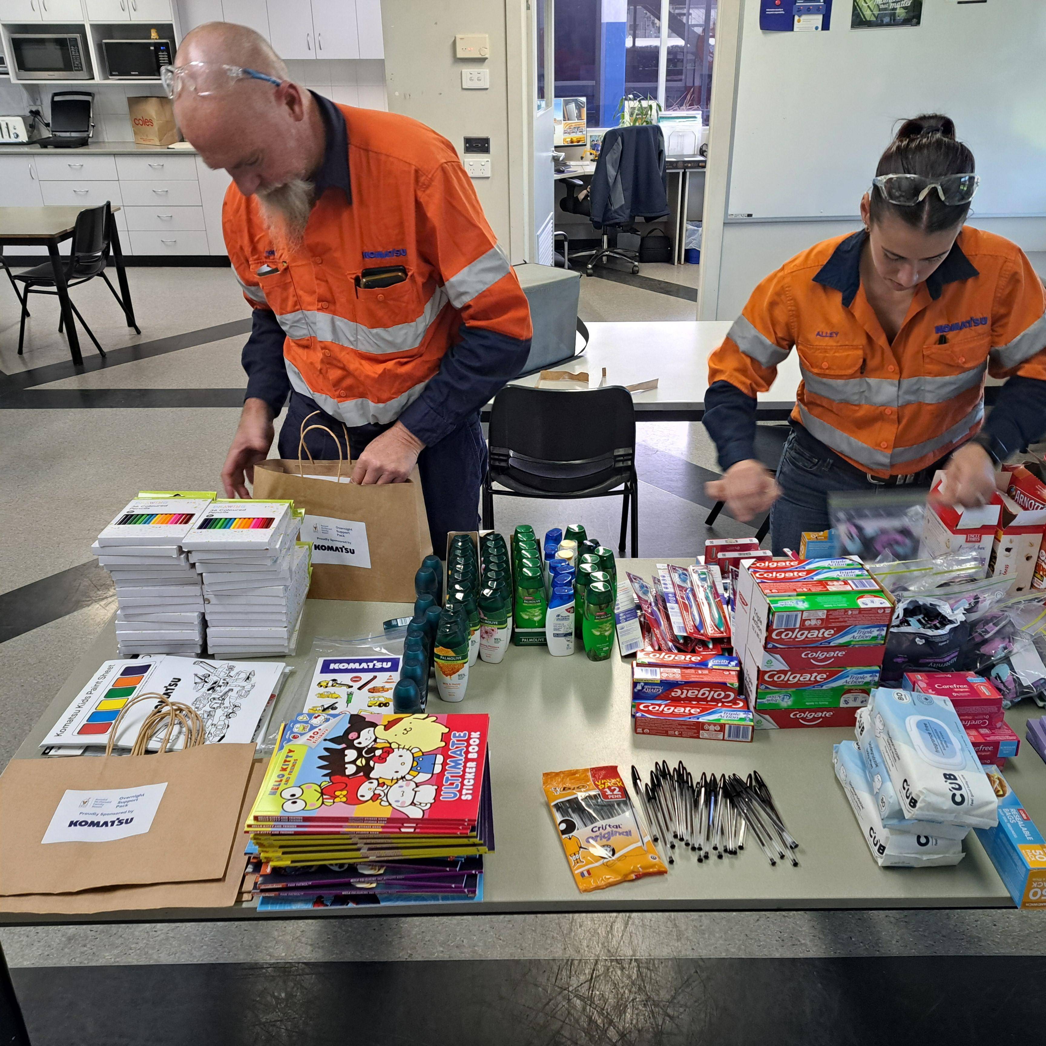 Two individuals in orange safety vests working at a table, organising various school supplies including notebooks, pens, and stationery items.