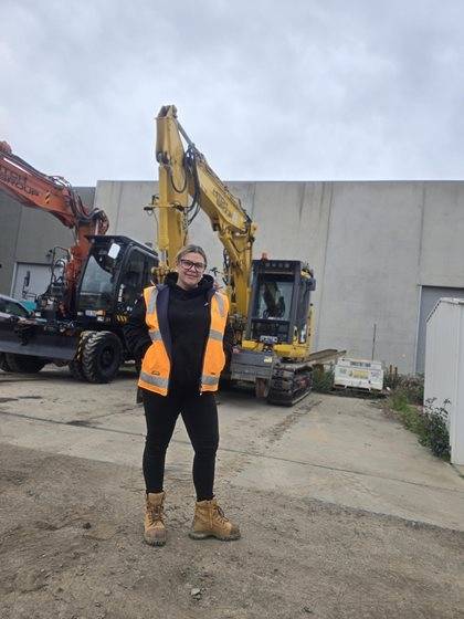 A person in a high-visibility vest and safety boots smiles in front of a yellow Komatsu excavator.
