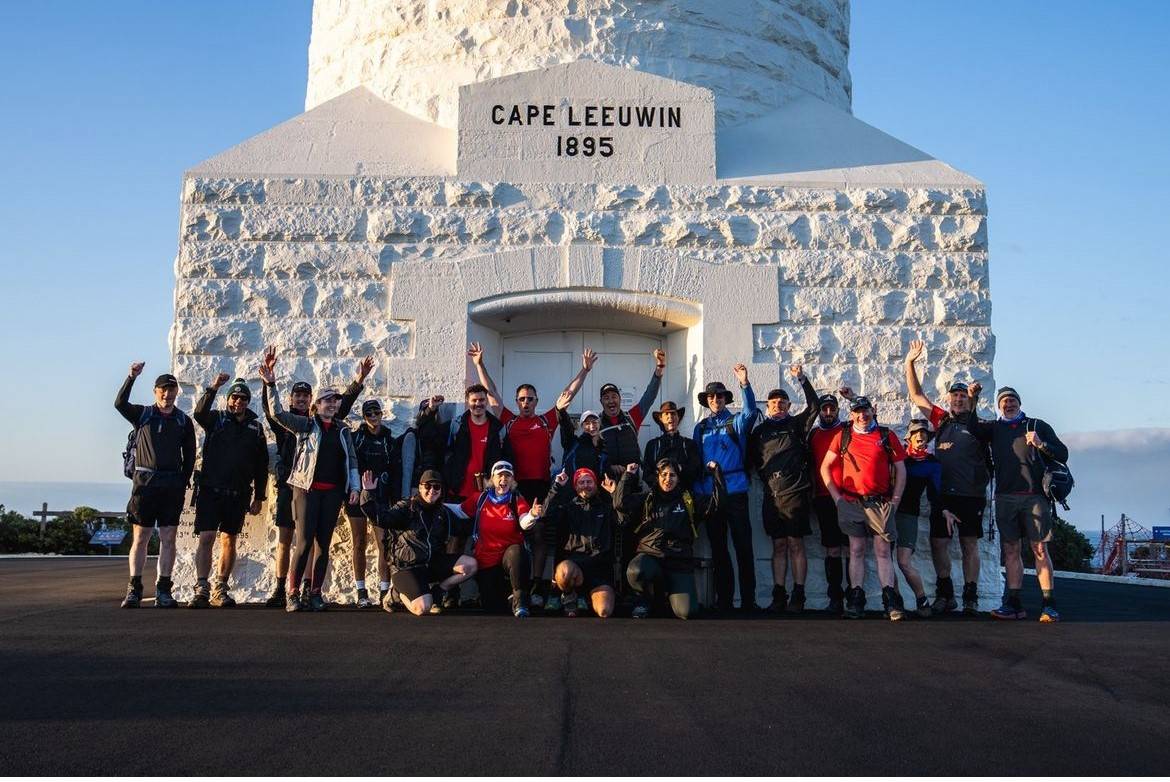 Group of people celebrating with raised arms in front of Cape Leeuwin Lighthouse, dated 1895, under a clear blue sky.