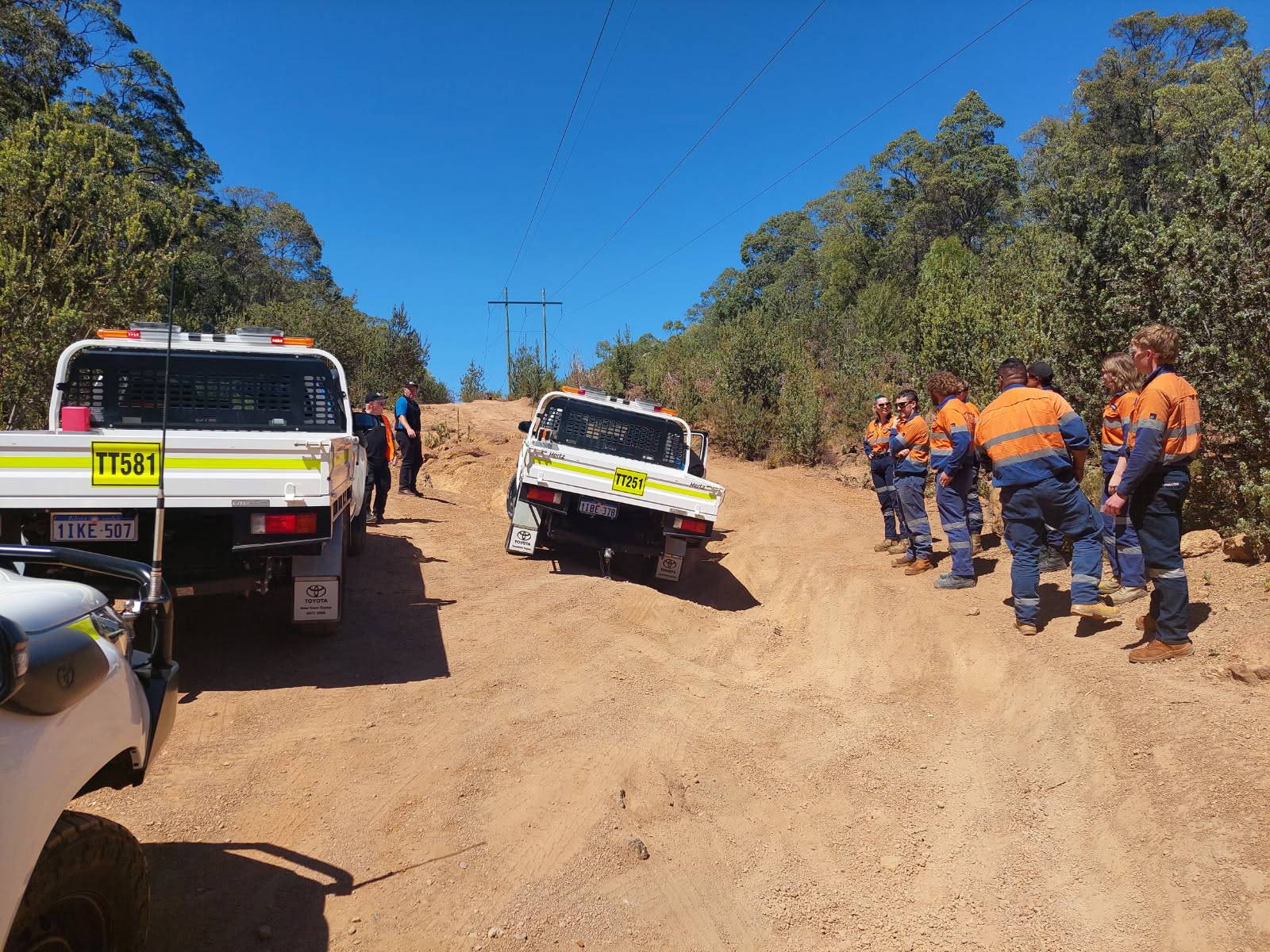 Individuals in high visibility workwear standing next to vehicles at a rural roadside scene, on a dirt road under a clear blue sky.