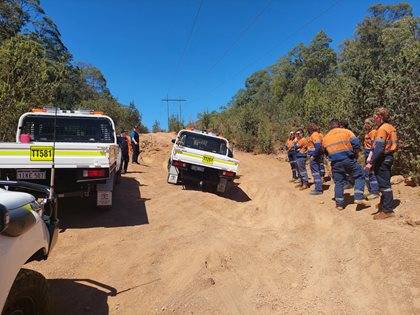 Individuals in high visibility workwear standing next to vehicles at a rural roadside scene, on a dirt road under a clear blue sky.