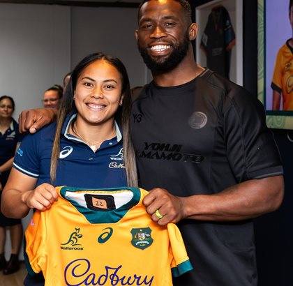 Two rugby players, one in a Wallaroos jersey and another in a black jersey, both smiling and holding up a Wallaroos jersey.