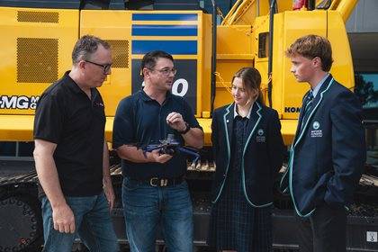 Three individuals listen attentively to another person holding a blue drone, standing in front of a large yellow Komatsu excavator.
