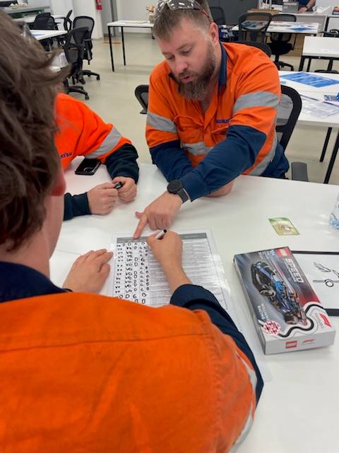 Two individuals in hi-vis vests are seated at a table, engaging in a discussion over a document with diagrams, in a classroom setting.