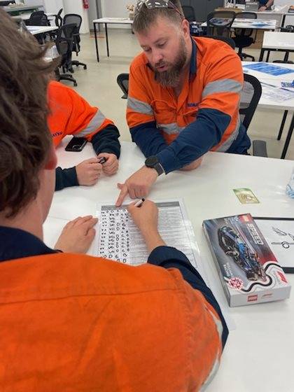 Two individuals in hi-vis vests are seated at a table, engaging in a discussion over a document with diagrams, in a classroom setting.
