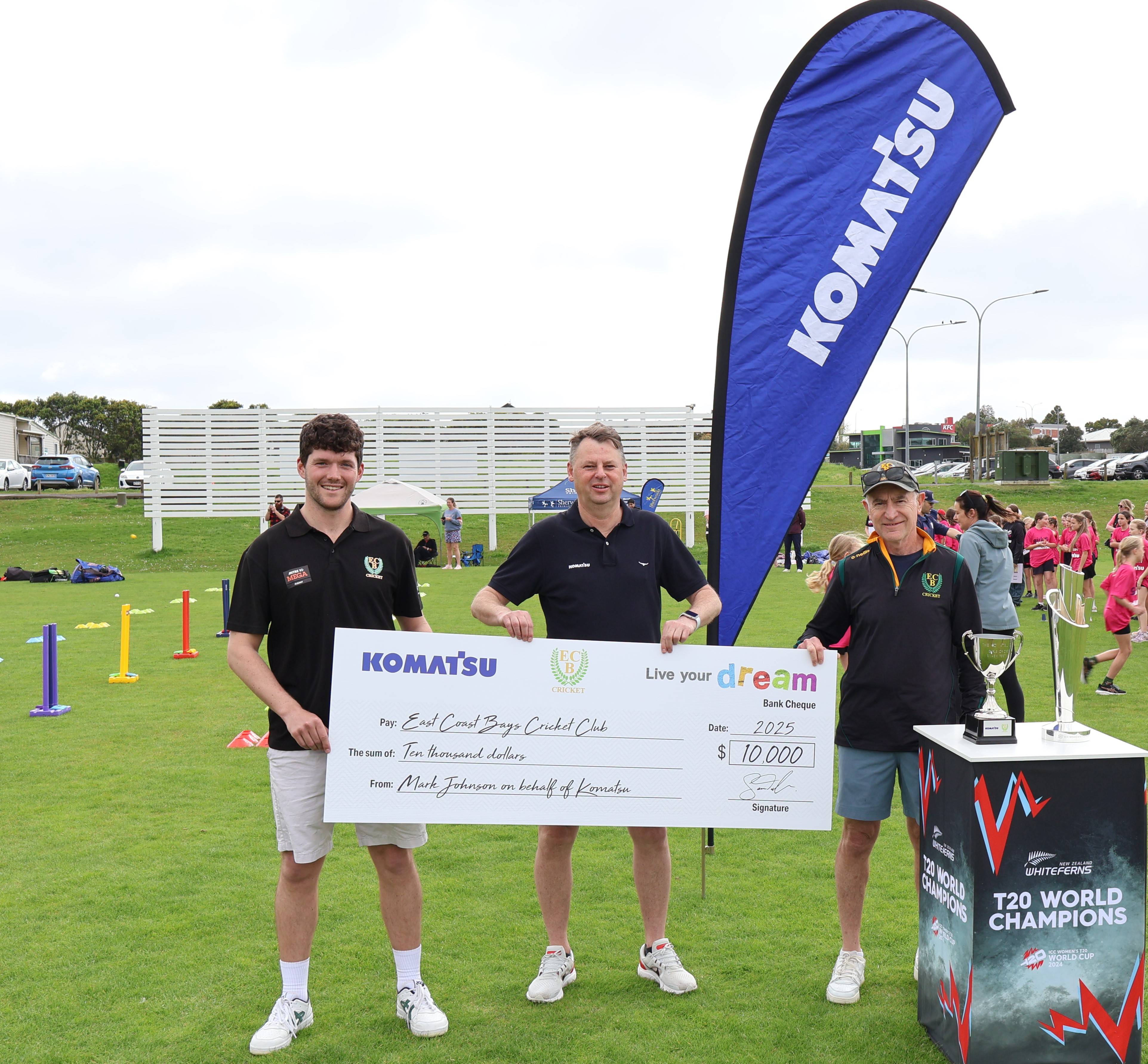 Three individuals posing outdoors with a large check for $10,000, presented by Komatsu. They are standing next to a Komatsu banner and a trophy, with sports equipment visible in the background.