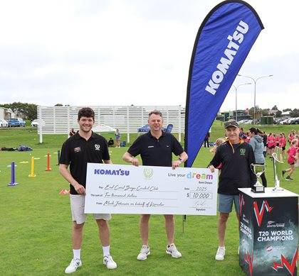 Three individuals posing outdoors with a large check for $10,000, presented by Komatsu. They are standing next to a Komatsu banner and a trophy, with sports equipment visible in the background.
