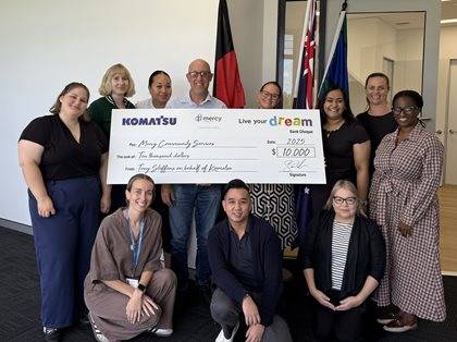 Group of people smiling and holding an oversized check from Komatsu to Mercy Community Services for $10,000 in an office setting with company banners in the background.