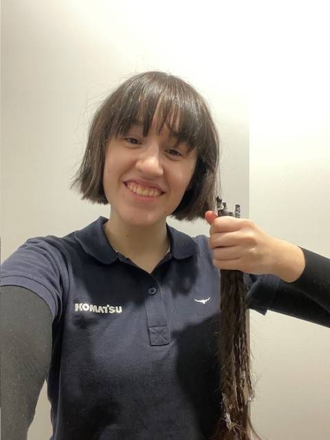 Person in a Komatsu polo shirt holding a long cut braid of hair and smiling at the camera.