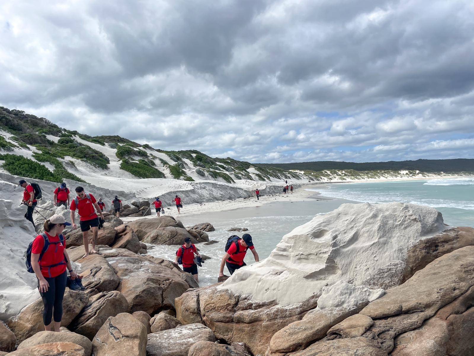 A group of people wearing red shirts exploring a rocky coastal area with white sand dunes under a cloudy sky.