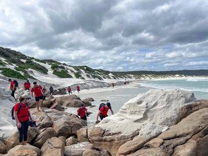 A group of people wearing red shirts exploring a rocky coastal area with white sand dunes under a cloudy sky.